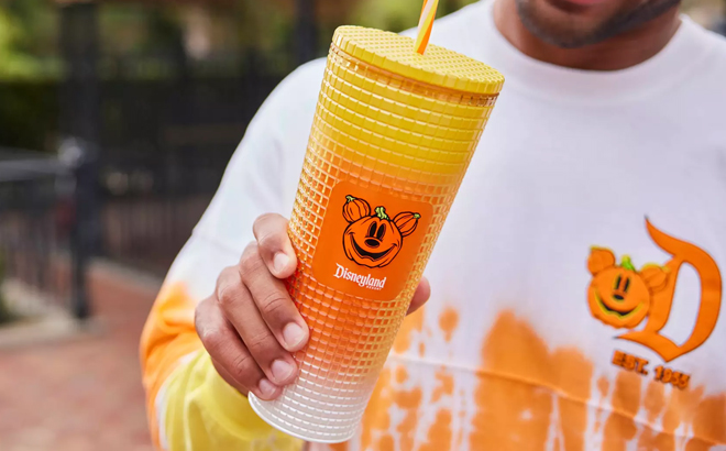 Someone in a Disneyland Halloween sweatshirt holding a textured orange-to-yellow tumbler with a Mickey pumpkin badge