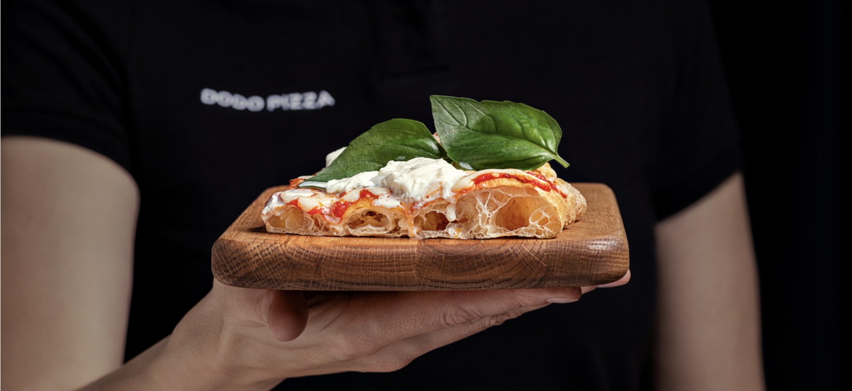 A hand holding a slice of pizza with burrata and basil on a wooden board, in front of a Dodo Pizza T-shirt
