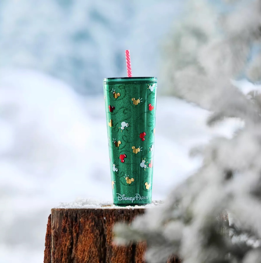 Green Disney Parks holiday tumbler with gold and red Mickey ornaments, standing on a snowy tree stump