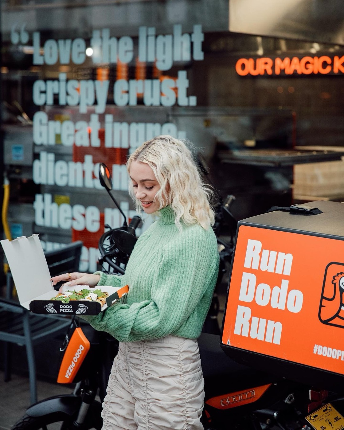 A customer opening a Dodo Pizza box beside a delivery scooter with a “Run Dodo Run” box, in front of the restaurant window
