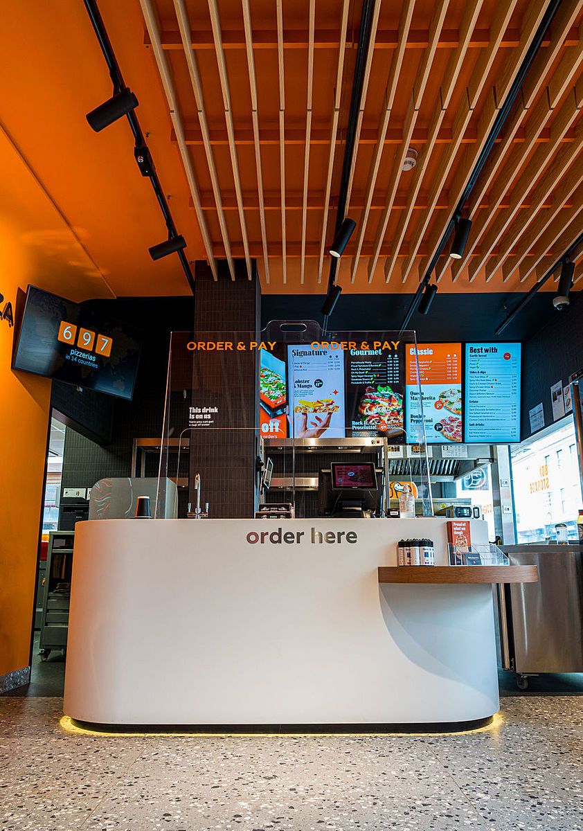 Order counter with “order here” lettering and digital menu screens above it, under an orange ceiling