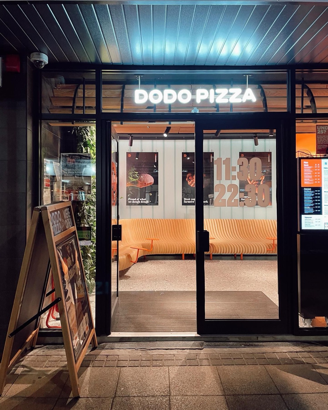 Dodo Pizza UK restaurant entrance at night, with an A-board and posters in the windows