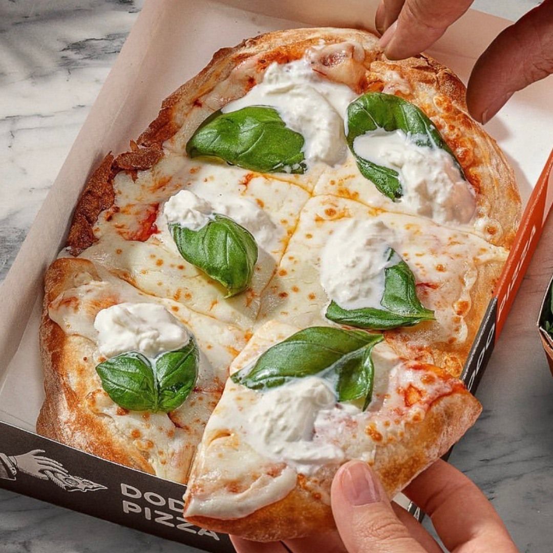 Hands taking a slice of burrata and basil pizza from a Dodo Pizza box on a marble table