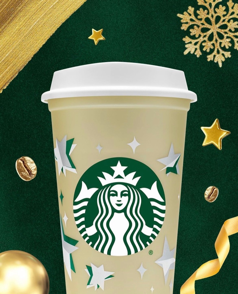Cream reusable hot cup with the Starbucks siren and white stars, on a green Christmas background with gold ribbon and coffee beans