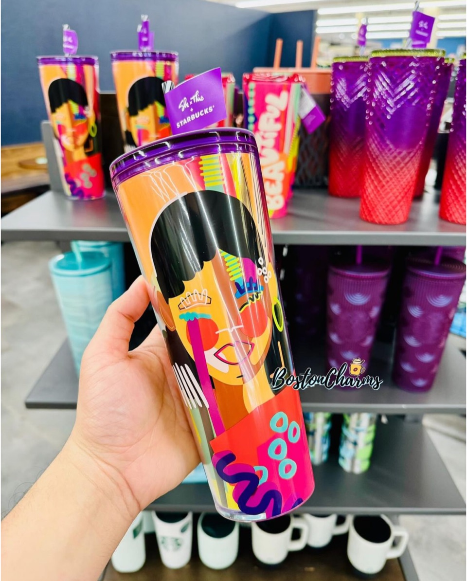A hand holding a “She Is This” cold cup with a portrait of a woman, in front of a store shelf